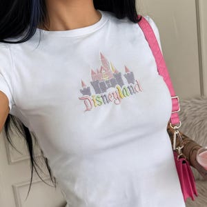 May include: White t-shirt with a colourful Disneyland castle design. The graphic features pink, yellow, and grey elements, with the word "Disneyland" in a rainbow font. The shirt has short sleeves.