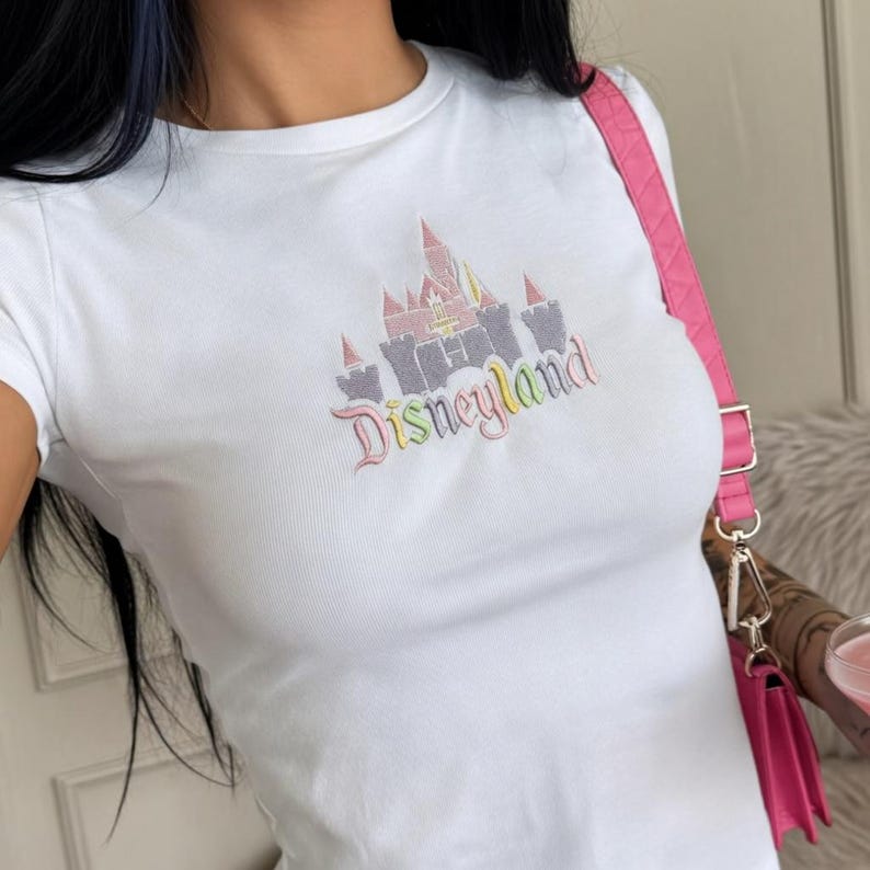 May include: White t-shirt with a colourful Disneyland castle design. The graphic features pink, yellow, and grey elements, with the word "Disneyland" in a rainbow font. The shirt has short sleeves.