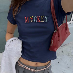 May include: A navy blue t-shirt embroidered with the word "Mickey" in various colours, featuring a cartoon mouse design. A red handbag with a zip is also visible.