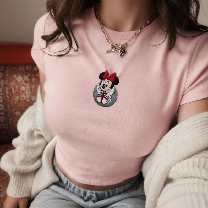May include: A light pink t-shirt featuring a Minnie Mouse patch with a red bow. The patch is circular and has a gray border. A gold chain necklace with a heart charm is also visible.