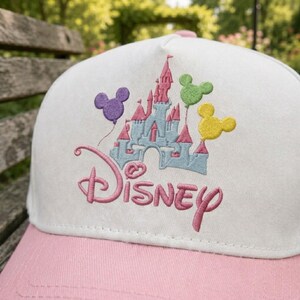 Puede incluir: Gorra de b&eacute;isbol blanca y rosa con un dise&ntilde;o bordado del castillo de Disney y globos de Mickey Mouse. La palabra "Disney" est&aacute; bordada en rosa. La gorra tiene una visera rosa y una corona blanca, perfecta para fans de Disney.