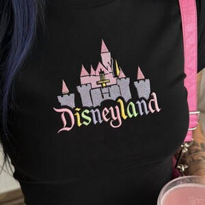 May include: Black t-shirt with an embroidered Disneyland castle design in pink, grey, and yellow. The word "Disneyland" is embroidered below the castle in a rainbow gradient. A pink strap is visible on the right side.