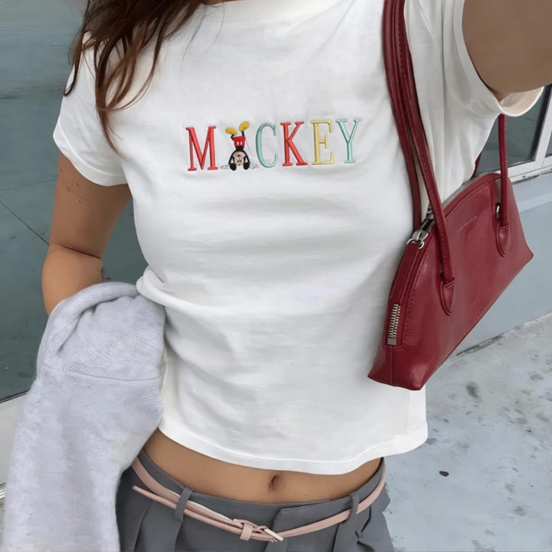 May include: White t-shirt embroidered with the word "Mickey" in colourful letters, with a cartoon character. A red handbag with a zip is visible. The person is wearing grey trousers and a pink belt.
