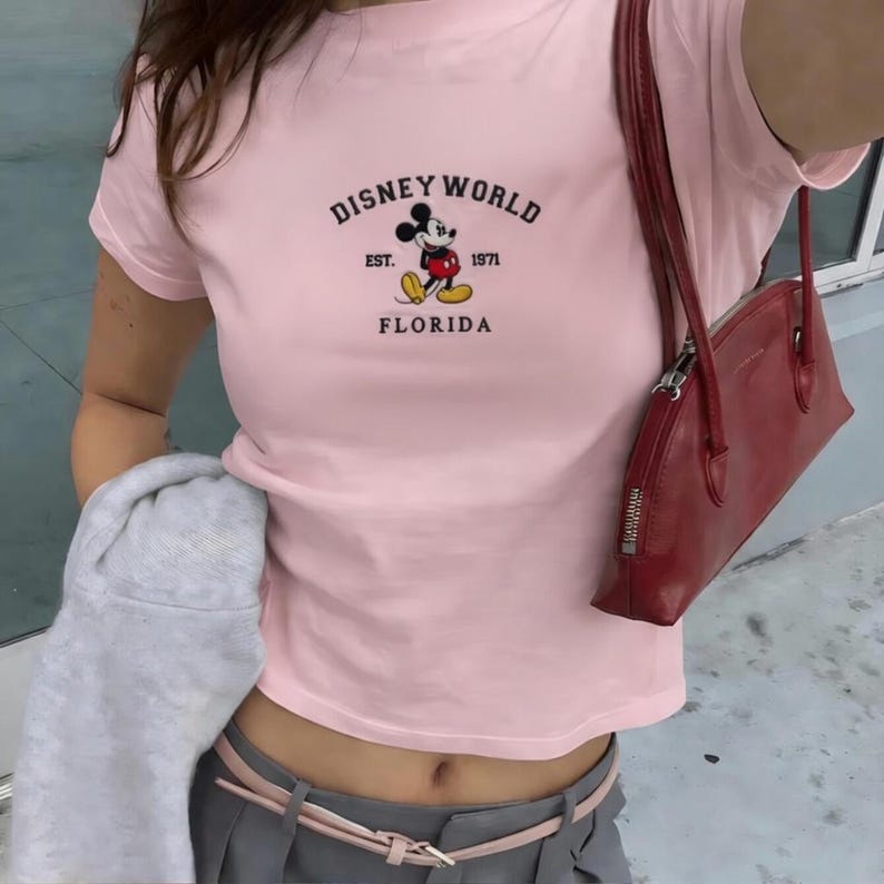 May include: A pale pink t-shirt featuring the text "DISNEY WORLD FLORIDA" and a Mickey Mouse graphic. The shirt has short sleeves and a crew neck. The graphic includes the text "EST. 1971".
