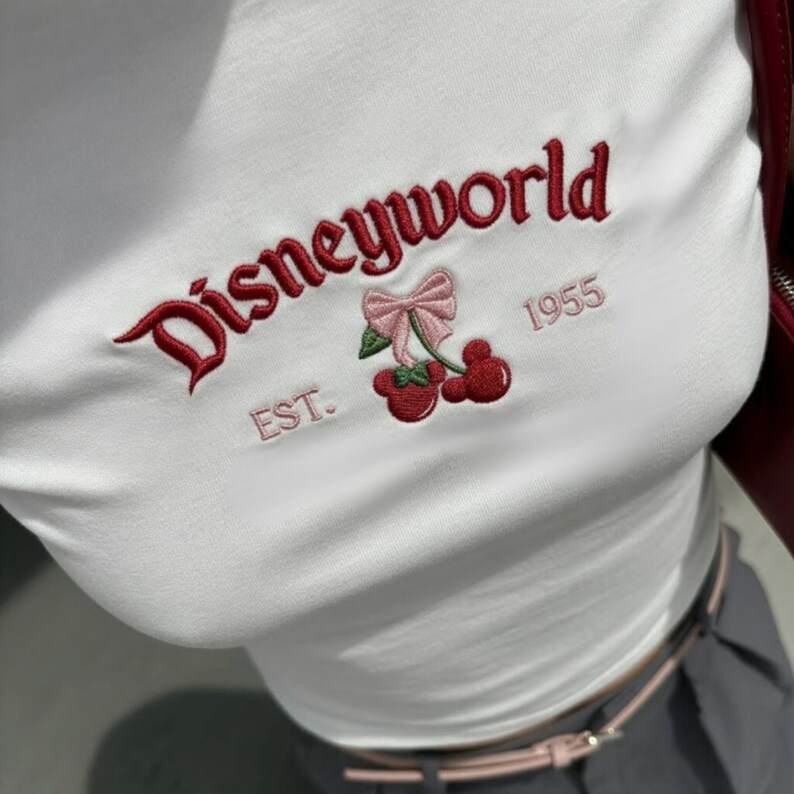 May include: White crop top with red embroidered "Disneyworld" text, a pink bow, and cherry design. The text "EST. 1955" is also embroidered on the shirt. The top is a fitted style.
