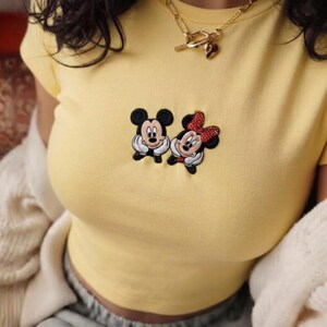 May include: A pale yellow short-sleeved t-shirt featuring a central embroidered graphic of Mickey and Minnie Mouse. The design shows both characters with their hands resting under their chins. A gold-toned chain necklace rests at the neckline.