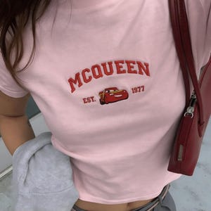 以下が含まれることがあります： ピンク色のTシャツ。赤色の刺繍で「McQUEEN」の文字、カートゥーンカーのグラフィック、そして「1977」の年号が描かれています。クルーネックと半袖のデザイン。右側には赤いハンドバッグが見えます。