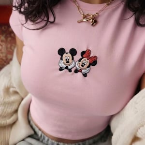 May include: A light pink short-sleeved t-shirt featuring a small embroidered graphic of Mickey and Minnie Mouse on the chest. The shirt is paired with a gold-toned chain necklace with a toggle clasp and a heart-shaped charm pendant.