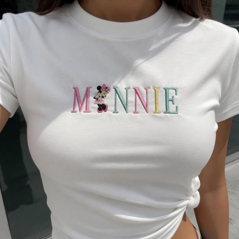 Puede incluir: Camiseta blanca con la palabra "MINNIE" bordada en colores pastel. La "M" es rosa, la "I" es verde, la "N" es rosa, la "N" es amarilla y la "E" es verde. Un dibujo de Minnie Mouse est&aacute; bordado encima de la "N".
