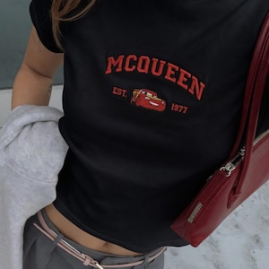以下が含まれることがあります： 赤い刺繍の「McQueen」の文字と漫画の車のグラフィックが特徴の黒いTシャツ。「EST. 1977」の文字がグラフィックの下にあります。赤いハンドバッグが画像の右側に表示されています。