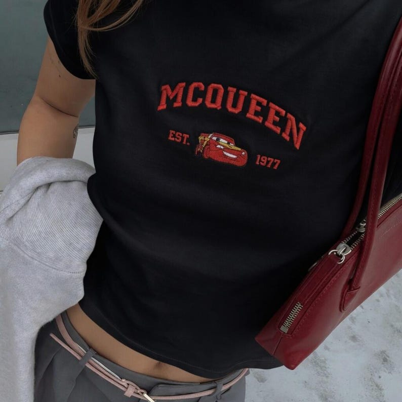 以下が含まれることがあります： 赤い刺繍の「McQueen」の文字と漫画の車のグラフィックが特徴の黒いTシャツ。「EST. 1977」の文字がグラフィックの下にあります。赤いハンドバッグが画像の右側に表示されています。