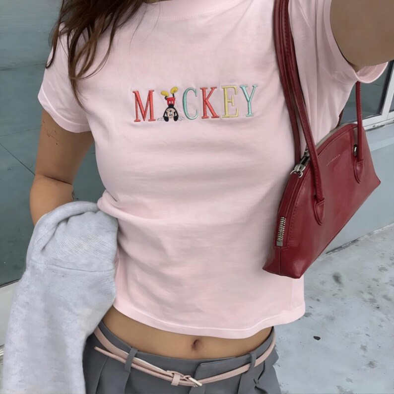 May include: A pale pink t-shirt featuring the embroidered word "Mickey" in various colours. The "M" and "C" are red, the "I" is black, the "K" is yellow, the "E" is blue, and the "Y" is green. A red handbag is visible.