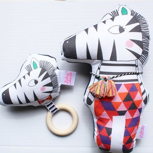 May include: Two black and white zebra plush toys. The larger zebra has a red, orange, and purple geometric pattern on its body. The smaller zebra has a wooden ring attached to it. Both zebras have the text "Julia Skye" on them.