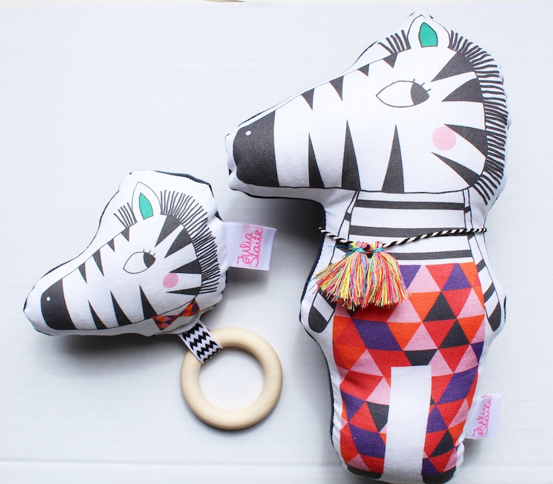 May include: Two black and white zebra plush toys. The larger zebra has a red, orange, and purple geometric pattern on its body. The smaller zebra has a wooden ring attached to it. Both zebras have the text "Julia Skye" on them.