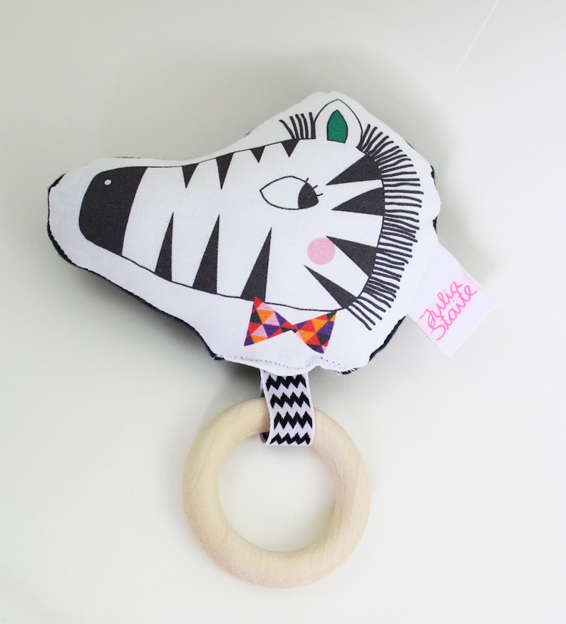 May include: A black and white zebra rattle with a colorful bow tie and a wooden ring. The rattle has a pink nose and green eyes. The tag says "Julia Staute".