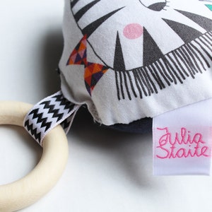 May include: A black and white zebra-patterned fabric toy with a pink circle and a green leaf. The toy has a wooden ring attached with a black and white zig-zag ribbon. The toy has a white label with the text "Julia Staite" in pink.
