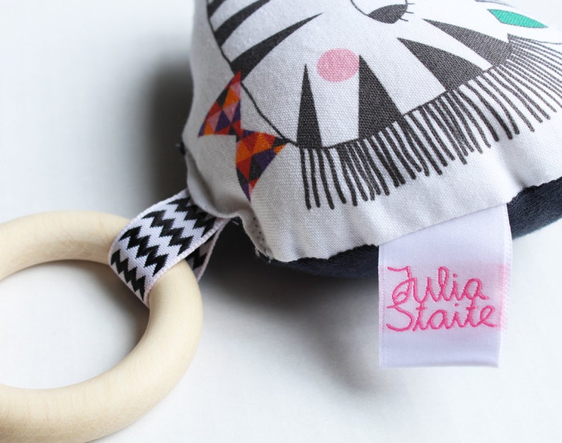 May include: A black and white zebra-patterned fabric toy with a pink circle and a green leaf. The toy has a wooden ring attached with a black and white zig-zag ribbon. The toy has a white label with the text "Julia Staite" in pink.