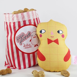 Peut inclure: Un jouet en peluche jaune en forme d'arachide avec un nœud papillon rouge et un visage amical. Le jouet est assis à côté d'un sac en papier rayé rouge et blanc contenant des cacahuètes. Le sac porte une étiquette qui dit "Pepe Peanut by Julia Staite".