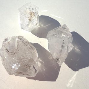 Peut inclure: Trois cristaux de quartz clairs aux formes naturelles sont présentés sur une surface blanche. Les cristaux ont des formes variées, avec des structures internes et des inclusions visibles. L'éclairage crée des ombres, soulignant les textures et les facettes des pierres.