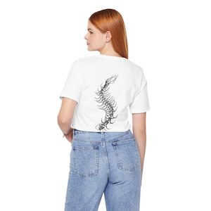 Può includere: Una t-shirt bianca a maniche corte con una dettagliata illustrazione grafica in bianco e nero di una colonna vertebrale umana che corre verticalmente lungo il centro della schiena. La t-shirt è abbinata a jeans in denim azzurro con due tasche.