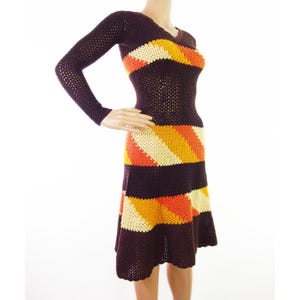 以下が含まれることがあります： 長袖で体にフィットするボディス、茶色、オレンジ、クリーム色のクロシェ編みドレス。ドレスはストライプ模様のフレアスカートです。