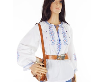 May include: White long-sleeve blouse with blue and red embroidery. The blouse has a round neckline and is paired with a brown leather belt with a silver buckle. A brown leather shoulder bag is also visible.