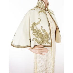 May include: Ivory cape with ornate gold embroidery. The cape features a high collar and a decorative border. The gold embroidery includes floral and paisley designs. The cape is worn over a cream-colored dress.