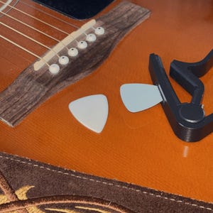 May include: Close-up of an acoustic guitar with a brown body and wooden neck. Two white guitar picks and a black capo are on the guitar's orange surface. The guitar's strings and bridge are visible.