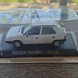 May include: A white Škoda Favorit 136 LS model car in a clear plastic display case. The model car is a small, detailed replica of the original vehicle. The text "ŠKODA FAVORIT 136 LS" is printed on the base.