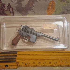 Könnte beinhalten: Ein silberfarbenes Miniatur-Pistolenmodell mit braunem Griff, eingeschlossen in einer transparenten Kunststoff-Blisterverpackung auf einer weißen Trägerkarte. Ein gelber Gliedermaßstab liegt davor und zeigt eine Länge von 16,5 cm.