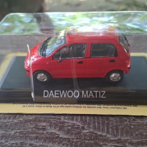 Könnte beinhalten: Ein rotes Daewoo Matiz Modellauto in einer durchsichtigen Kunststoff-Präsentationsbox. Das Fahrzeug hat schwarze Türgriffe, Seitenspiegel und silberfarbene Räder. Der schwarze Sockel ist mit dem weißen Schriftzug DAEWOO MATIZ versehen.