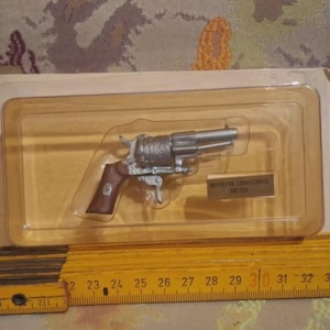 May include: A silver-colored toy revolver with a brown handle encased in a clear plastic blister pack on a cardboard backing. A yellow folding ruler is positioned in front, showing the toy measures approximately 5.5 inches in length.