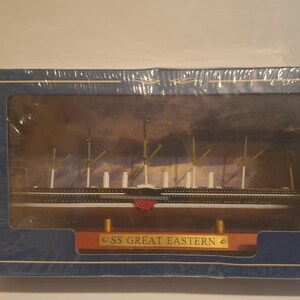 Op de afbeelding: Een model van het schip SS Great Eastern in een blauwe doos. Het scheepsmodel is wit, zwart en goud, met verschillende masten en een rode accentkleur. De doos heeft een helder venster en de tekst "SS GREAT EASTERN".
