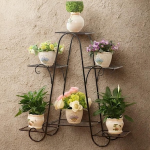 Może przedstawiać: Czarny, kutego żelaza stojak na rośliny z wieloma półkami, na których znajdują się różne rośliny doniczkowe i kwiaty. Stojak ma łukowaty kształt i jest zamontowany na teksturowanej ścianie. Doniczki są kremowe z kwiatowymi wzorami.