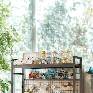 Puede incluir: Un escritorio moderno con una estantería de dos niveles, con figuras decorativas, una pequeña lámpara y un espejo. El escritorio es blanco, con un organizador de rejilla y varios artículos de papelería. El fondo muestra una gran ventana con vistas a la vegetación.