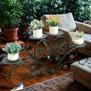 May include: A dark green metal plant stand with four small, round platforms. Each platform holds a white pot with colorful flowers. The stand has a heart-shaped design with leaf accents, ideal for indoor plants.