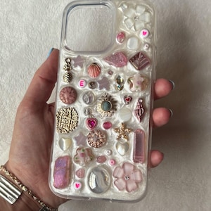 May include: Clear phone case decorated with various charms, including pink flowers, hearts, stars, and a gold-colored charm with the letter "E". The case has a pearlescent background and is embellished with beads and other decorative elements.