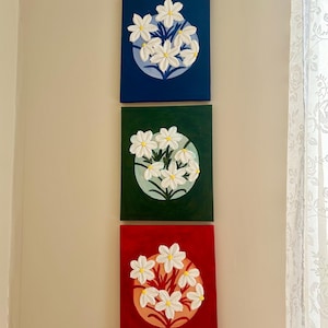 May include: Three square canvas paintings featuring white flowers with yellow centres. Each painting has a different background colour: navy blue, dark green, and red. The flowers are arranged in a circular pattern.