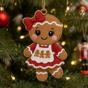 May include: A gingerbread person ornament featuring brown embroidery, a red bow, and a red dress with a white apron. The apron displays three small gingerbread figures. The ornament has a gold hanging loop and a smiling face with pink cheeks.