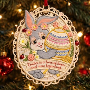 May include: Embroidered Easter decoration with a grey bunny holding a decorated egg. The bunny wears a floral crown and is surrounded by flowers and ladybirds. The ornament has a scalloped edge and a ribbon for hanging. Text reads "Easter is a time of hope and new beginnings."