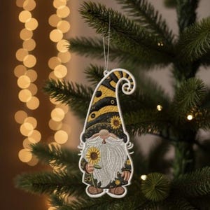 May include: Embroidered gnome Christmas ornament with a black and yellow striped hat and a white beard. The gnome is holding a sunflower. The ornament is hanging on a Christmas tree with lights in the background.