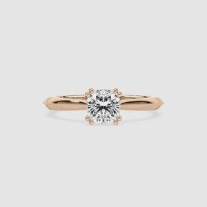 May include: A rose gold engagement ring with a square-cut diamond center stone. The band features a delicate beaded design and tapers towards the sides. The diamond is held in place by four prongs.