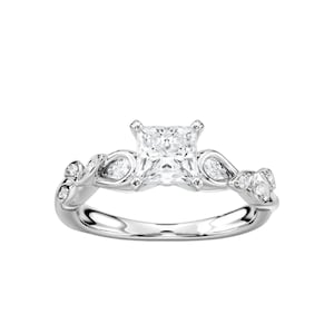 Pode incluir: Um anel de noivado prateado com uma pedra central com corte princesa. A banda apresenta um design floral com pequenas pedras de destaque. O anel está sobre um fundo branco, realçando os seus detalhes intrincados e brilho.