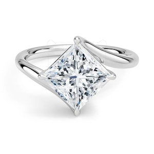 May include: A silver ring featuring a large, square-cut diamond. The diamond is set in a prong setting and has a brilliant, multifaceted appearance. The ring's band curves gracefully around the finger, creating a modern and elegant design.