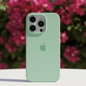 May include: A light green phone case with a triple-lens camera cutout and the Apple logo. The case is made of a smooth material and is set against a backdrop of pink flowers and a blue sky.