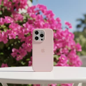 May include: A light pink phone case with a triple-lens camera and the Apple logo. The case is on a white table with a vibrant pink flowering bush in the background. The sky is blue.