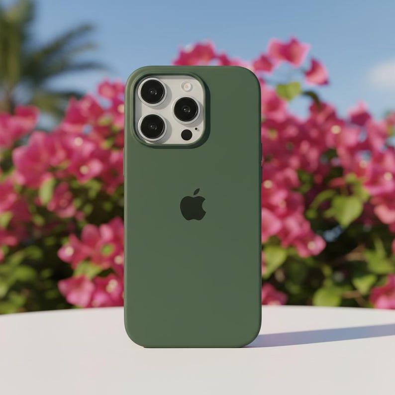 May include: A dark green phone case with the Apple logo. The case has a smooth finish and covers the back and sides of the phone. The camera lenses are visible at the top. The background features pink flowers and a blue sky.