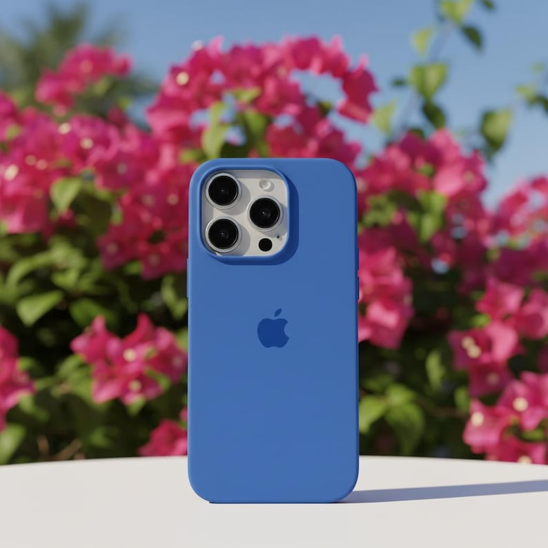 May include: A blue phone case with the Apple logo. The case has a smooth finish and a camera cutout. The background features pink flowers and green foliage under a blue sky.