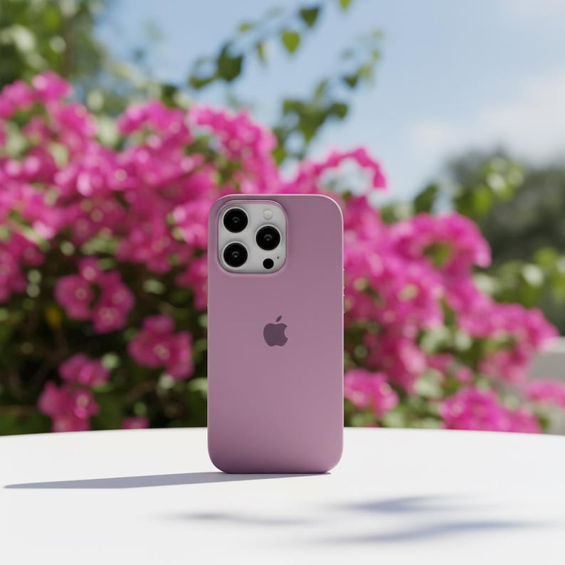 May include: A mauve-colored phone case with a triple-lens camera cutout and the Apple logo. The case is on a white surface, with a backdrop of vibrant pink flowers and a clear blue sky.