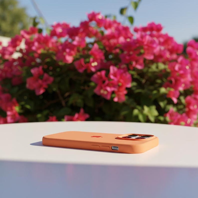 May include: A peach-colored phone case with a camera cutout and the Apple logo. The case is lying on a white surface, with a backdrop of vibrant pink flowers and green foliage.